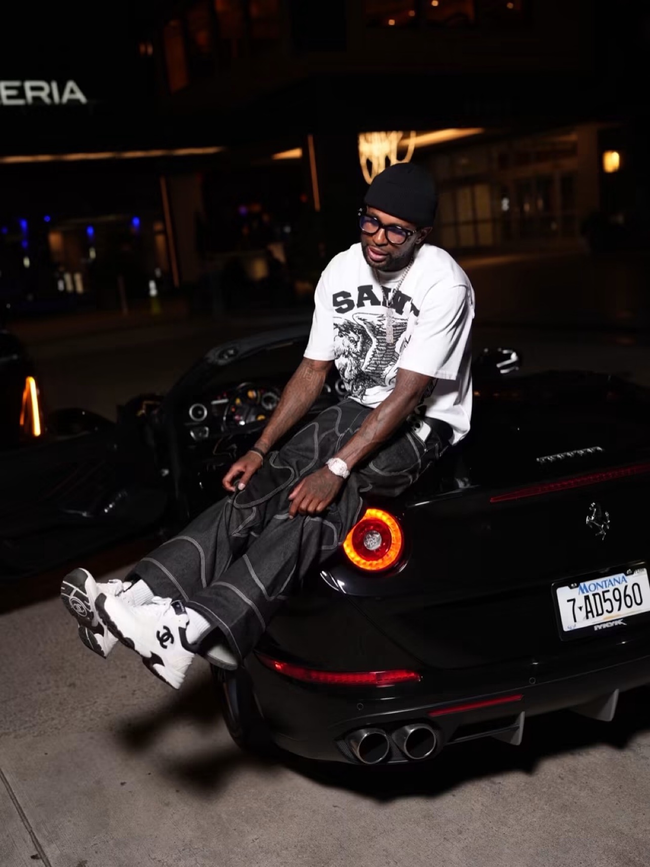 Bossman Yayo sitting on the rear of a black Ferrari at night, legs over the taillight