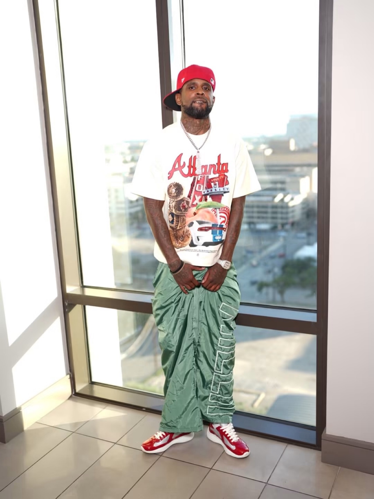 Bossman Yayo standing by a high-rise window, wearing a red cap and an Atlanta graphic tee